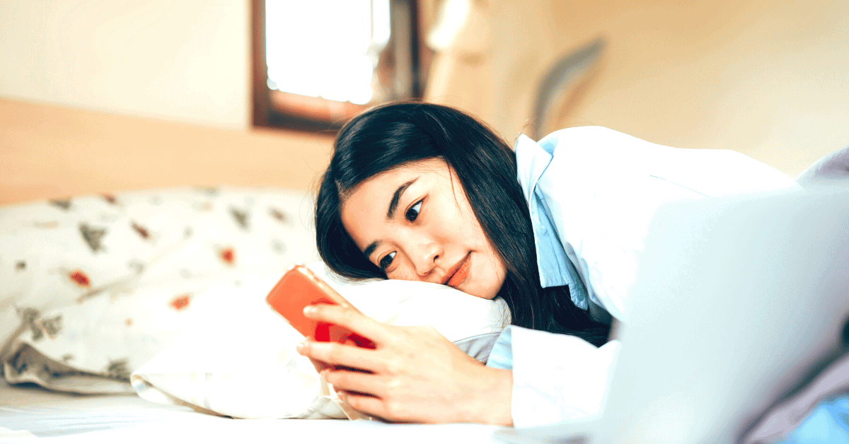 A young woman lays on her bed and looks at her phone