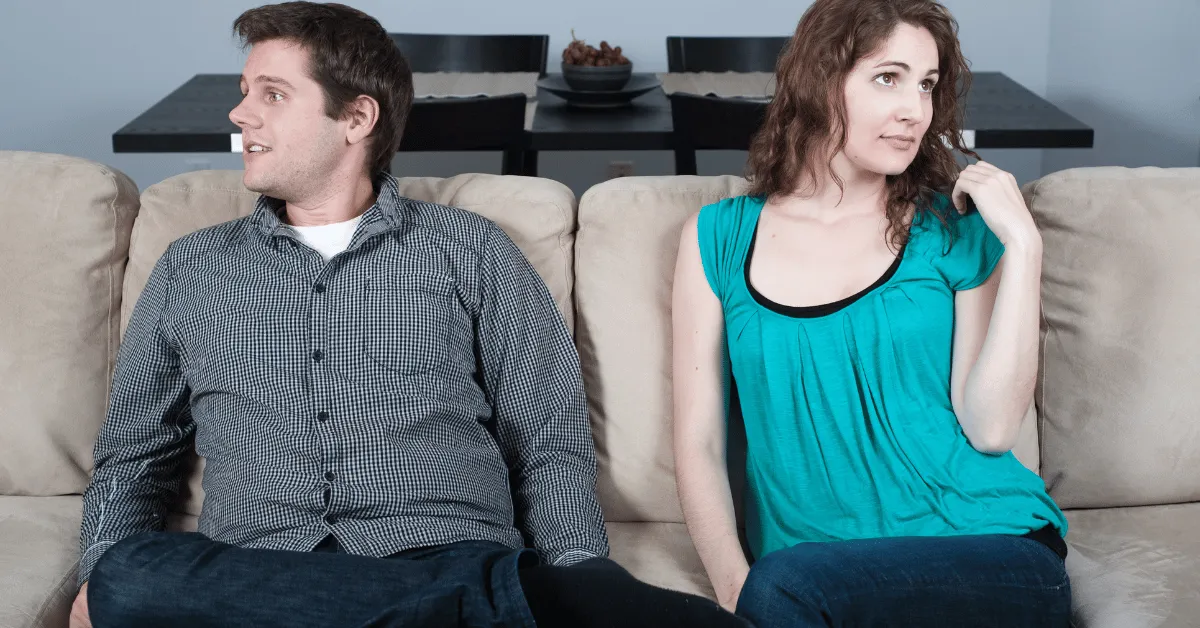 A couple sits on a couch leaning away from each other looking uncomfortable